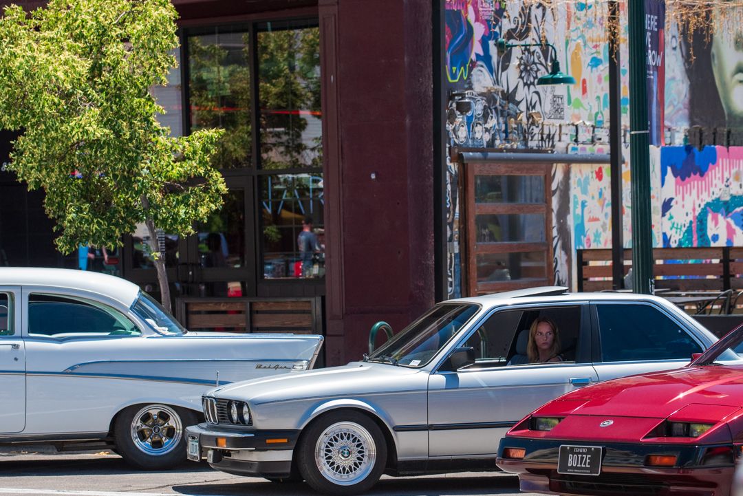 A woman in a silver BMW E30 stopped in downtown Boise traffic between a classic Chevrolet Bel Air and a red Toyota MR2, in front of a colorful street mural, with an Idaho vanity plate reading BOIZE visible