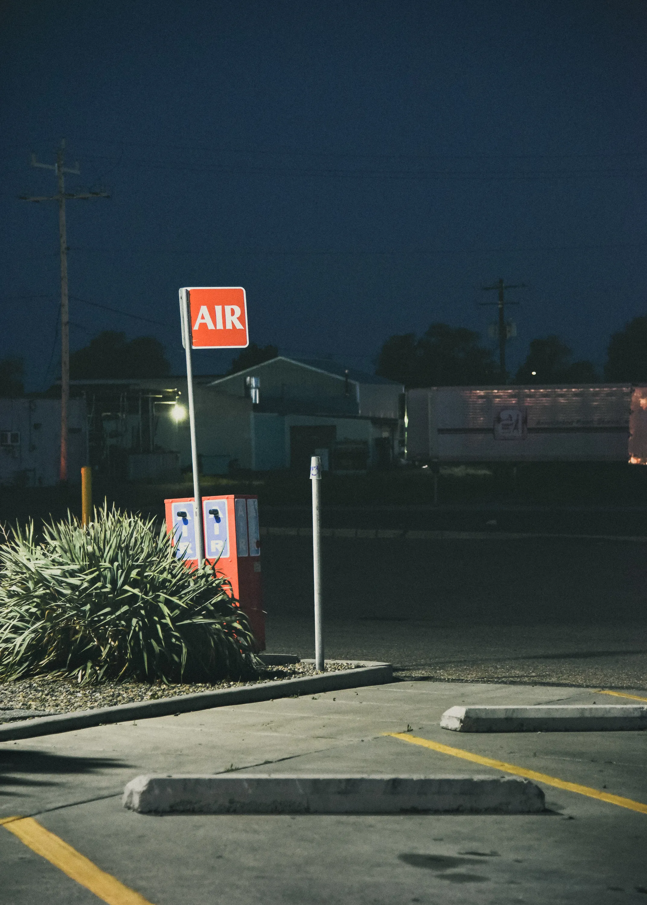 A red AIR sign on a pole at a gas station at night, beside an air pump machine partially obscured by shrubs, with an empty parking lot in the foreground and dark industrial buildings in the background