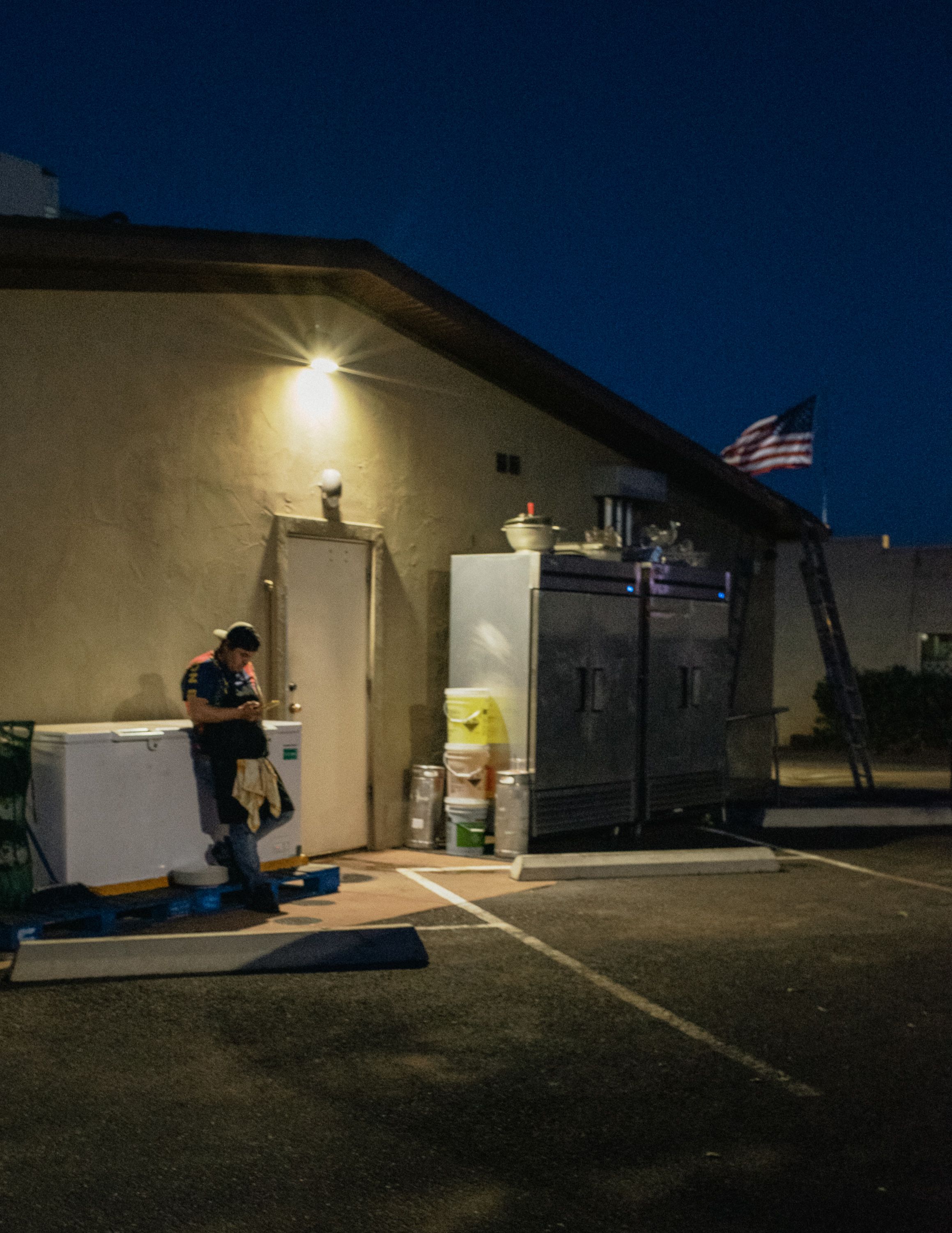 A Hispanic back of house worker takes a break behind a restaurant; Lit by a single overhead light, scrolling on his phone with an American flag in the background.