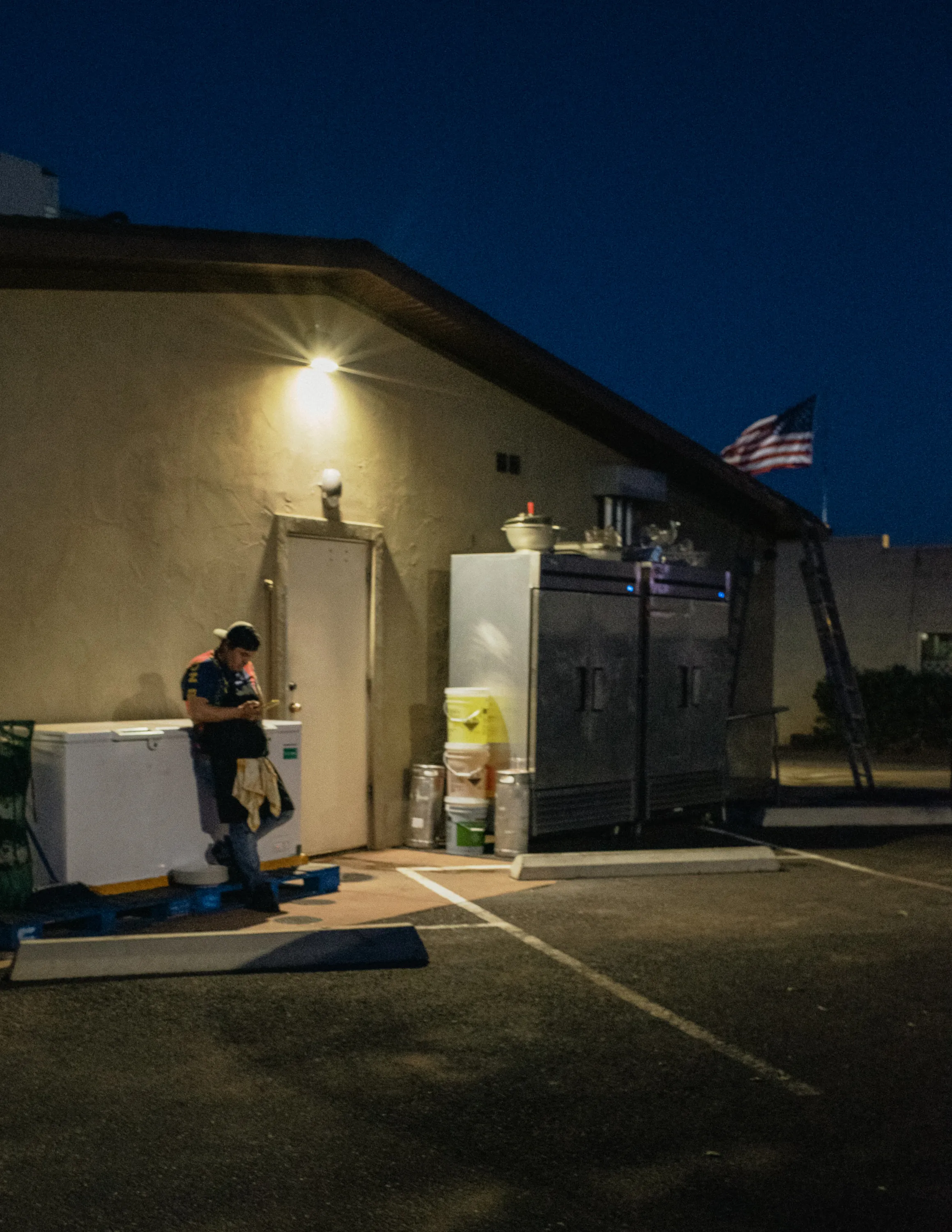 A Hispanic back of house worker takes a break behind a restaurant; Lit by a single overhead light, scrolling on his phone with an American flag in the background.