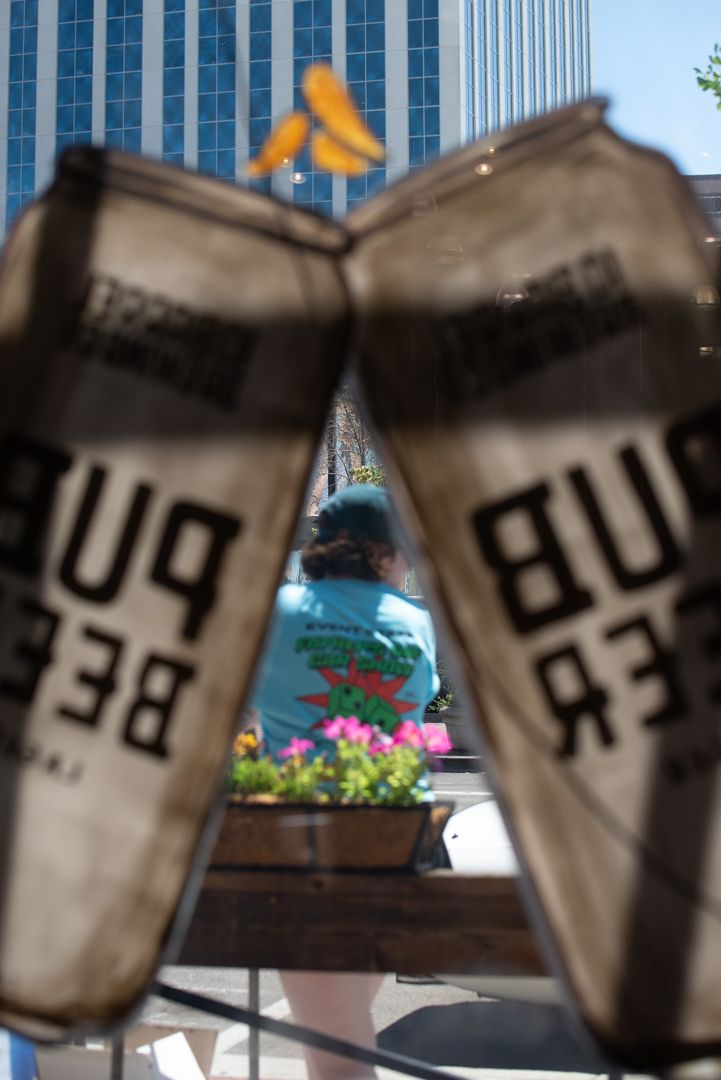 A man in a graphic t-shirt seen through reversed hand-painted Pub Beer can signage at 10 Barrel Brewing Co, with a downtown Boise office tower visible behind him