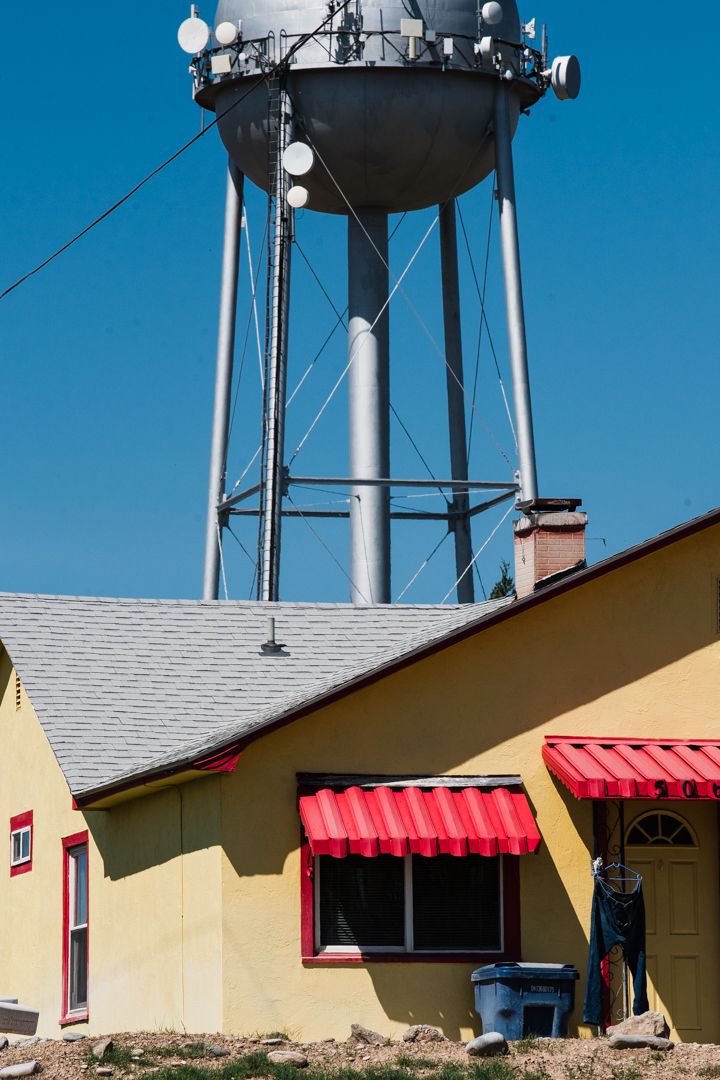 A yellow house with red awnings and jeans hanging beside the door, with a municipal water tower covered in cellular equipment looming behind it in Notus, Idaho