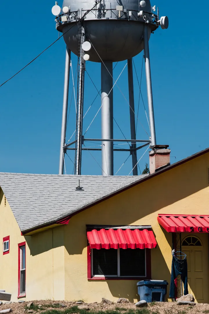 A yellow house with red awnings and jeans hanging beside the door, with a municipal water tower covered in cellular equipment looming behind it in Notus, Idaho