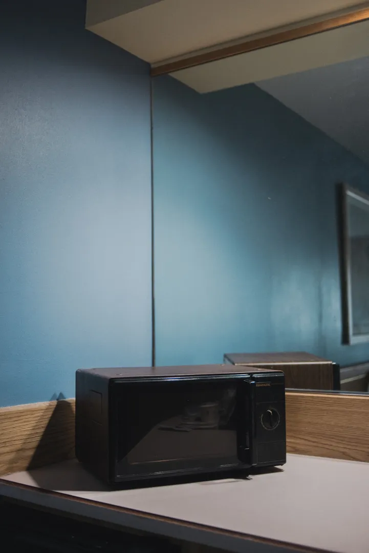 A black microwave on a dresser in a budget motel room in Kanab, Utah; its geometric lines echoed by the mirror edge, wall corner, and angular shadows against blue walls
