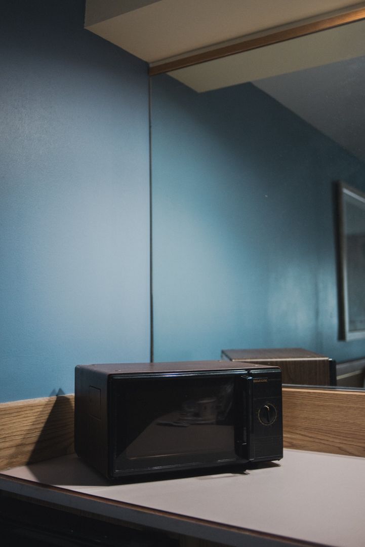 A black microwave on a dresser in a budget motel room in Kanab, Utah; its geometric lines echoed by the mirror edge, wall corner, and angular shadows against blue walls