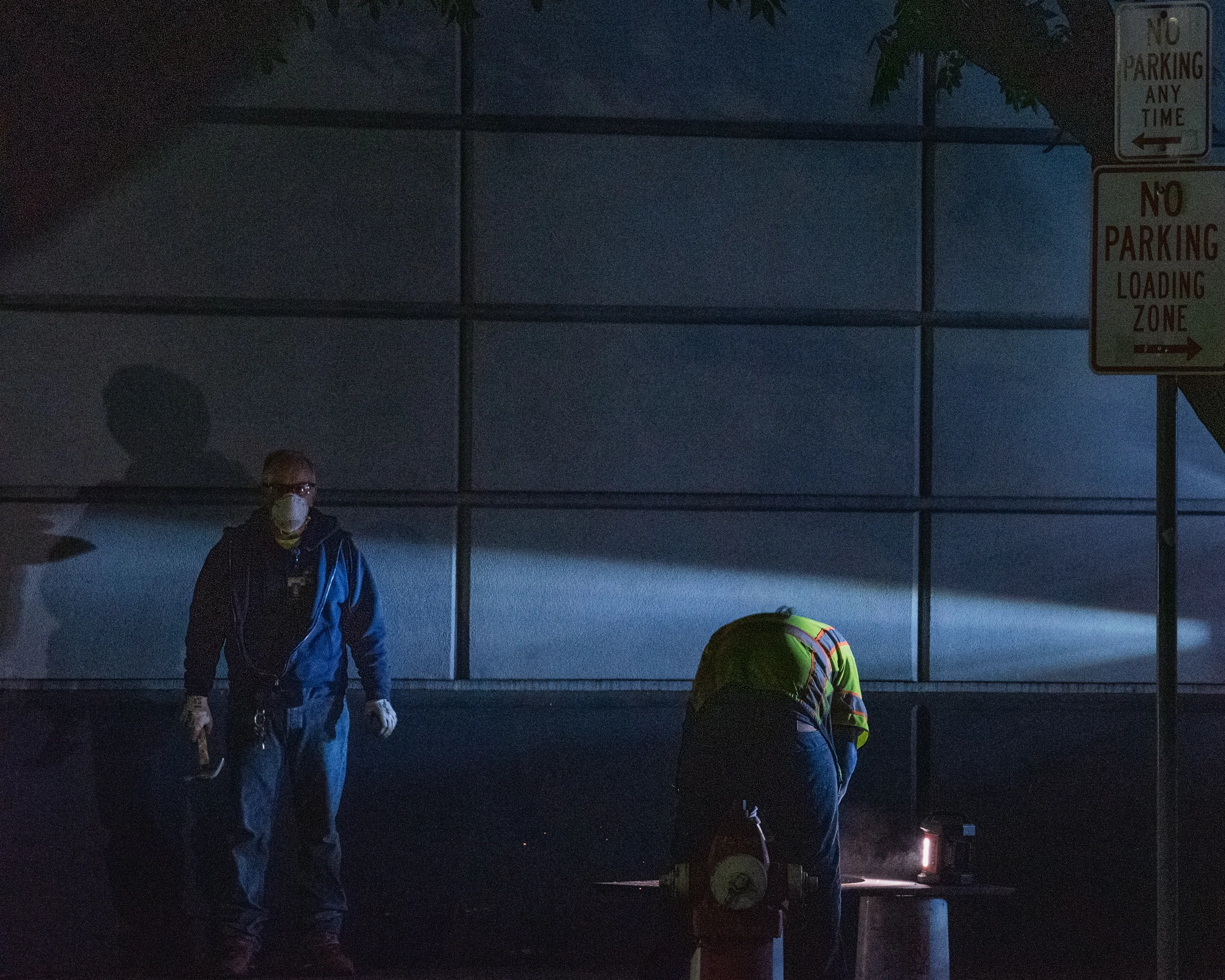 Two workers in a downtown Boise loading zone before dawn, one in a yellow safety vest using an angle grinder while the other stares intensely into the camera, masked, with a hammer in hand